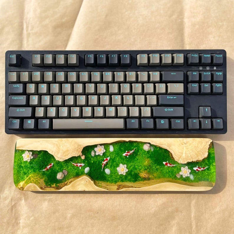Puede incluir: Un teclado mec&aacute;nico negro con teclas grises y azul claro se encuentra encima de un reposamu&ntilde;ecas de madera. El reposamu&ntilde;ecas presenta un dise&ntilde;o de resina verde y dorada con peces koi, flores blancas y peque&ntilde;as rocas.