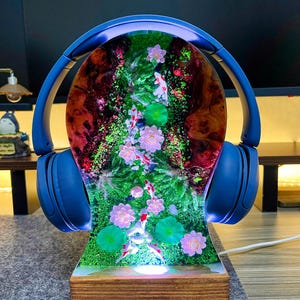 May include: A blue headphone stand with a resin design featuring koi fish, pink flowers, and green lily pads. The stand has a wooden base and is illuminated with a soft light. The headphones are blue.