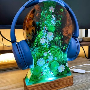 May include: A blue pair of headphones rests on a decorative headphone stand. The stand features a resin design with a koi pond scene, including fish, flowers, and lily pads. The base is made of wood, and the stand is illuminated with a soft glow.