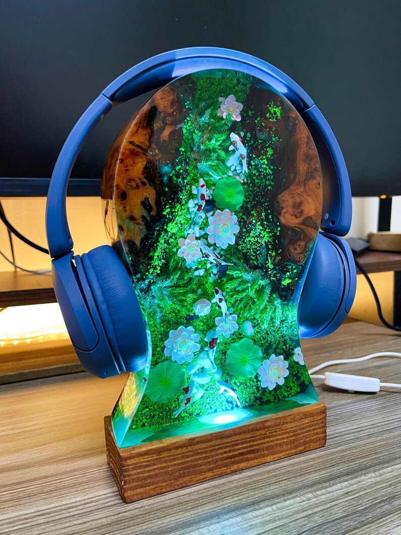 May include: A blue pair of headphones rests on a decorative headphone stand. The stand features a resin design with a koi pond scene, including fish, flowers, and lily pads. The base is made of wood, and the stand is illuminated with a soft glow.