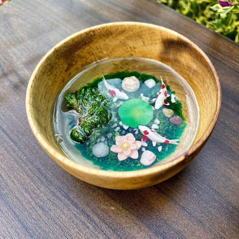 May include: A round wooden bowl containing a clear resin pour that mimics a pond. Inside the resin are three small white and red koi fish, a bright green lily pad, a pale pink lotus flower, green moss-like textures, and small white pebbles.
