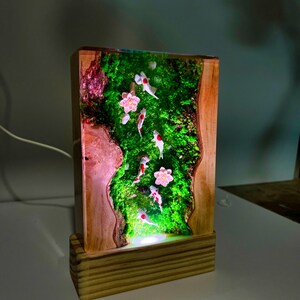 Puede incluir: L&aacute;mpara rectangular de madera con un panel de resina que representa una escena de estanque de carpas koi en miniatura. El panel muestra peces koi nadando en un entorno verde musgoso con flores rosas. La l&aacute;mpara tiene una base de madera y una luz c&aacute;lida.