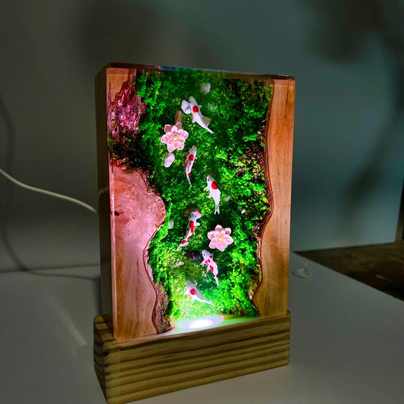 Puede incluir: L&aacute;mpara rectangular de madera con un panel de resina que representa una escena de estanque de carpas koi en miniatura. El panel muestra peces koi nadando en un entorno verde musgoso con flores rosas. La l&aacute;mpara tiene una base de madera y una luz c&aacute;lida.