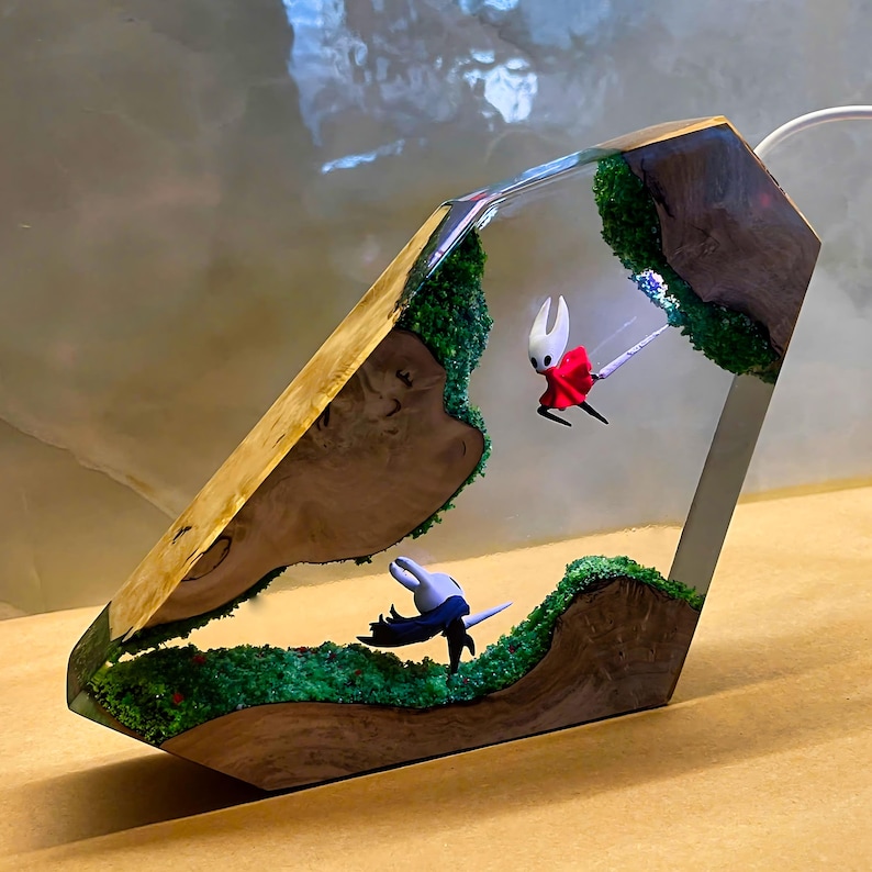 May include: A unique, geometric resin lamp featuring two small character figurines set within a landscape of green moss and wood. The lamp's exterior is a mix of wood and clear resin, with a warm, inviting glow.