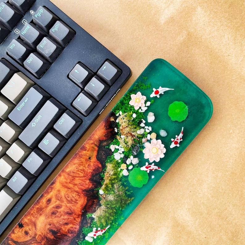 May include: A black mechanical keyboard next to a custom resin wrist rest. The wrist rest features a vibrant design with koi fish, flowers, and a wood-like pattern. The resin is a mix of green and brown.