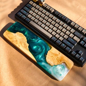 May include: A wooden keyboard wrist rest with a green and blue epoxy resin inlay. The resin has a swirling pattern and resembles water. The wrist rest is sitting on top of a black mechanical keyboard with white keycaps.