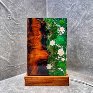May include: A rectangular lamp with a wooden base. The lamp's body features a wood grain design on one side and a pond scene with koi fish, lily pads, and flowers on the other. The lamp is lit with a soft glow.