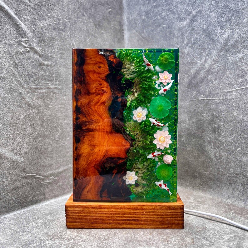 May include: A rectangular lamp with a wooden base. The lamp's body features a wood grain design on one side and a pond scene with koi fish, lily pads, and flowers on the other. The lamp is lit with a soft glow.
