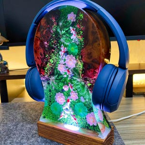May include: A unique headphone stand featuring a resin design with koi fish, flowers, and greenery. The stand is set on a wooden base and has blue headphones resting on top. The resin design is illuminated with a soft glow.