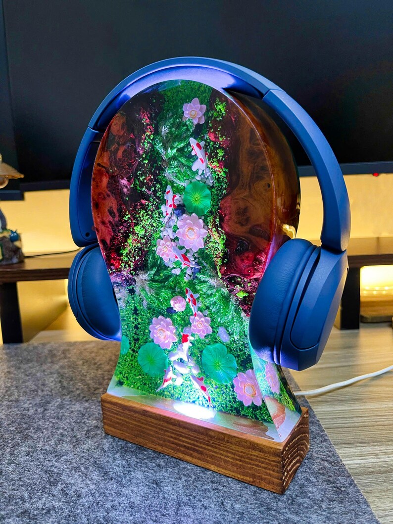 May include: A unique headphone stand featuring a resin design with koi fish, flowers, and greenery. The stand is set on a wooden base and has blue headphones resting on top. The resin design is illuminated with a soft glow.