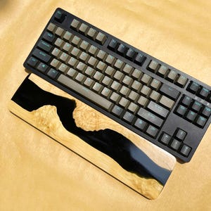 K&ouml;nnte beinhalten: Eine schwarze mechanische Tastatur mit beigefarbenen Tastenkappen und einer Holz-Handballenauflage. Die Handballenauflage hat ein schwarzes Harz-Fluss-Design mit nat&uuml;rlicher Holzmaserung. Die Tastatur hat ein Standard-Layout.