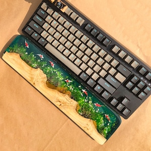 May include: A wooden keyboard wrist rest with a green and blue epoxy resin top featuring a koi fish design. The wrist rest is paired with a black mechanical keyboard with white keycaps and blue backlighting.