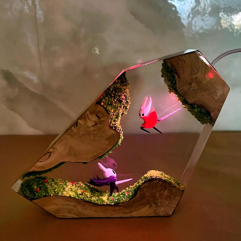 May include: A unique resin lamp featuring two animated characters in a diorama setting. The lamp has a wooden base with moss and floral accents, and the characters are in mid-action. The lamp is illuminated with a warm, red glow.
