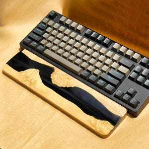 K&ouml;nnte beinhalten: Eine schwarze mechanische Tastatur mit hellbraunen Tastenkappen und einer h&ouml;lzernen Handgelenkauflage. Die Handgelenkauflage hat ein Design aus schwarzem Epoxidharz in hellfarbenem Holz. Die Tastatur hat schwarze Tasten und einen schwarzen Rahmen.