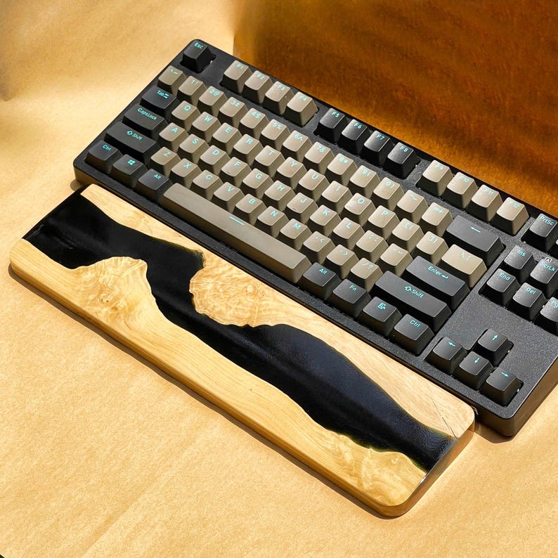 K&ouml;nnte beinhalten: Eine schwarze mechanische Tastatur mit hellbraunen Tastenkappen und einer h&ouml;lzernen Handgelenkauflage. Die Handgelenkauflage hat ein Design aus schwarzem Epoxidharz in hellfarbenem Holz. Die Tastatur hat schwarze Tasten und einen schwarzen Rahmen.