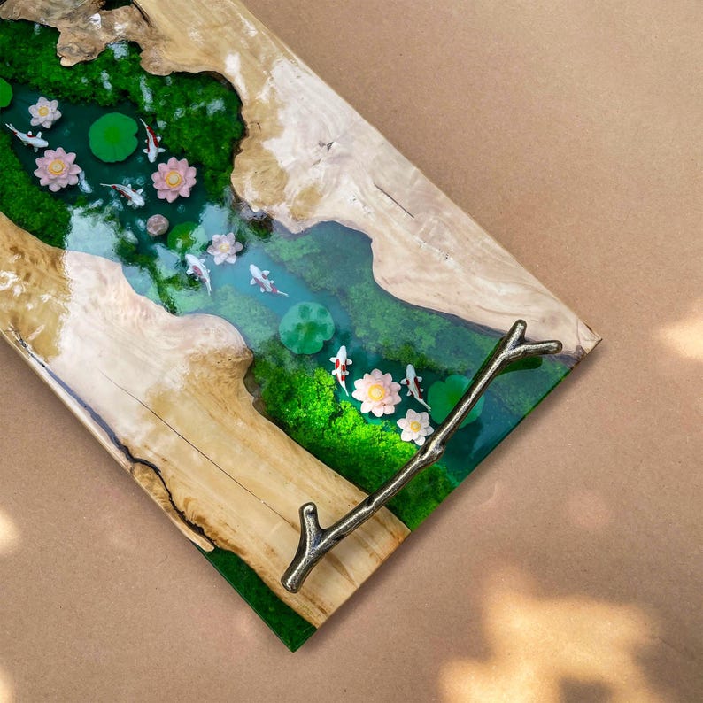 May include: A decorative wooden serving tray with a river scene. The tray features a clear epoxy river with koi fish, lily pads, and flowers. The tray has a natural wood edge and a metal branch handle.