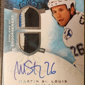 May include: A hockey trading card featuring Martin St. Louis of the Tampa Bay Lightning. The card is blue and silver with a silver emblem of a lightning bolt. The card has a piece of St. Louis' jersey attached to it. The card is signed by St. Louis.