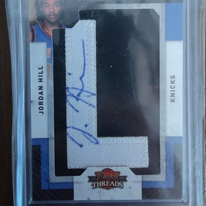 May include: A white jersey patch with a blue signature and the text "ROOKIE CLASS" and "KNICKS" on a black background. The patch is framed in a clear plastic case with the text "Panini THREADS" on the bottom.