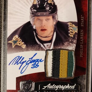 May include: A hockey card featuring Philip Larsen of the Dallas Stars. The card is a rookie card and features an autograph of the player. The card is in a plastic case.