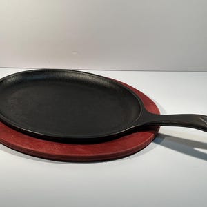 May include: A black cast iron oval-shaped skillet with a wooden base. The skillet has a handle and is sitting on a red wooden base.