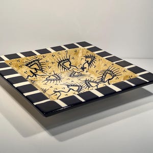 May include: Square ceramic decorative tray with a black and white chequered border. The centre of the tray is beige with black abstract figures. A unique piece of home decor.