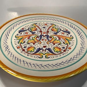 May include: A large, round, white ceramic serving platter with a yellow rim. The platter features a colorful floral design in shades of blue, green, yellow, orange, and red.