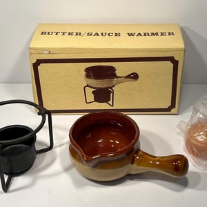 May include: A brown ceramic butter or sauce warmer with a handle. The warmer is on a black metal stand with a black metal base. The warmer is in its original box, which says "Butter/Sauce Warmer".