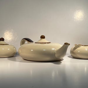 May include: A cream-coloured ceramic teapot with brown trim, a matching creamer, and a sugar bowl.