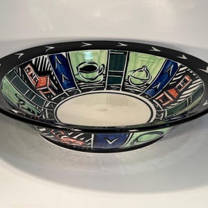 May include: A ceramic bowl with a black rim and a white interior. The bowl has a colourful interior design with green, blue, and orange geometric shapes, and illustrations of a coffee cup and sugar bowl. The rim has white arrow-shaped designs.