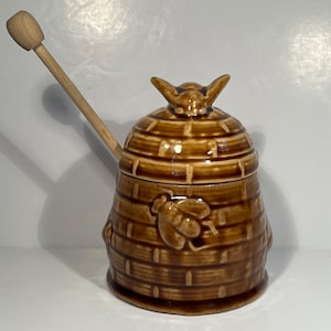 Op de afbeelding: Een bruine keramische honingpot in de vorm van een bijenkorf met een houten roerstaaf. De pot heeft een bijenontwerp op de deksel en de zijkant.