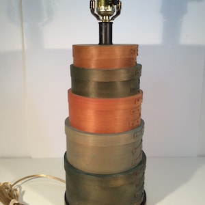 May include: A table lamp made of stacked wooden boxes painted in shades of brown, green, and orange. The lamp has a black metal base and a gold cord.