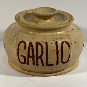 Könnte beinhalten: Ein beiger Knoblauchtopf aus Keramik mit Deckel. Das Wort "GARLIC" ist in dunkelbraunen Buchstaben auf der Vorderseite geschrieben. Der Topf hat ein gesprenkeltes Aussehen und einen kleinen Knopf auf dem Deckel.