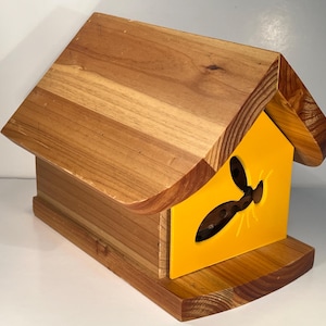 May include: A wooden bee house with a yellow front panel featuring a cutout of a bee. The house has a pitched roof and a small landing platform.