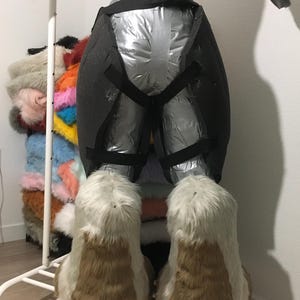 May include: A partially constructed fursuit form, wrapped in silver material and black straps. The feet are large, with white and brown faux fur. A rack of colorful faux fur scraps is visible in the background.