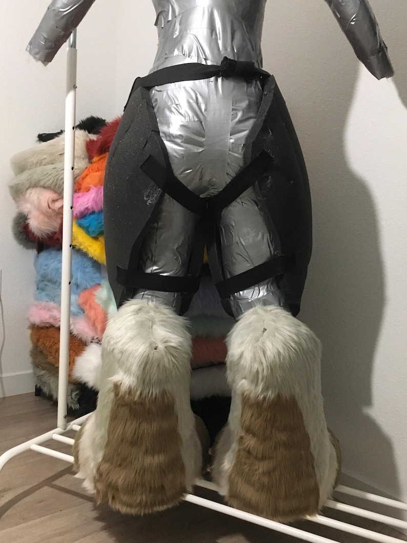 May include: A partially constructed fursuit form, wrapped in silver material and black straps. The feet are large, with white and brown faux fur. A rack of colorful faux fur scraps is visible in the background.