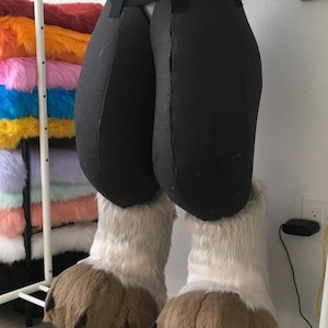 May include: A partially constructed fursuit featuring black padded pants and large, fluffy, light brown and white paws. The torso is covered in silver tape. A rack of colorful faux fur is visible in the background.