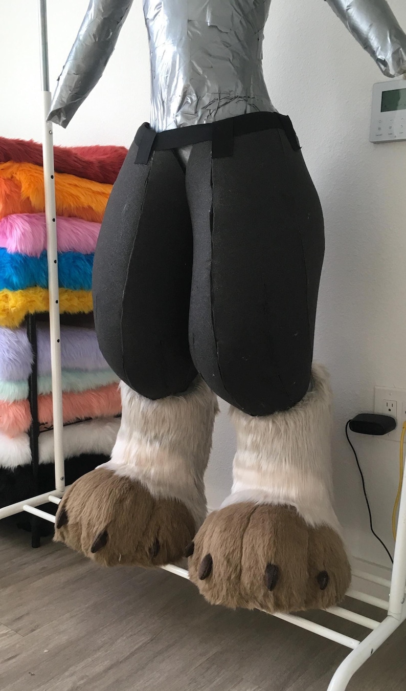 May include: A partially constructed fursuit featuring black padded pants and large, fluffy, light brown and white paws. The torso is covered in silver tape. A rack of colorful faux fur is visible in the background.