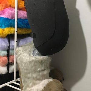 May include: A black foam leg and a furry foot of a costume. The foot is brown and white with brown claws. A rack of colorful faux fur fabric is in the background.