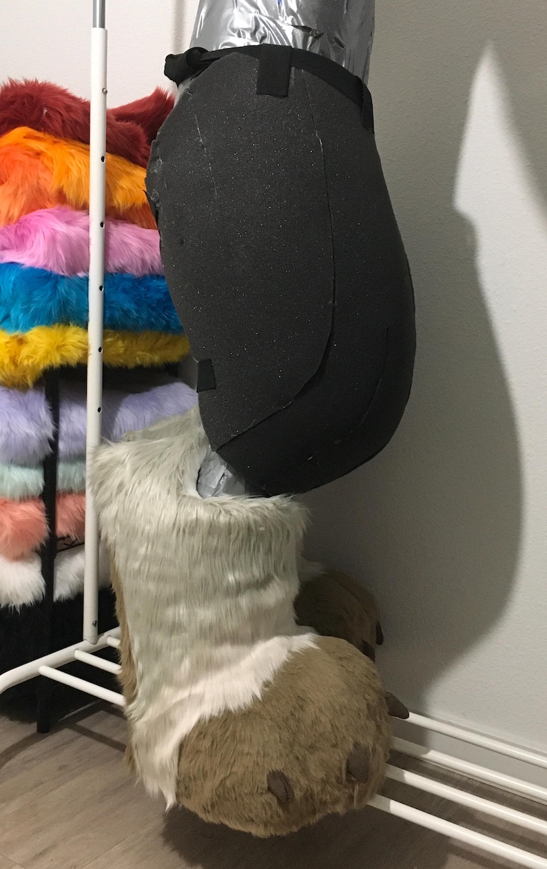 May include: A black foam leg and a furry foot of a costume. The foot is brown and white with brown claws. A rack of colorful faux fur fabric is in the background.