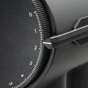 May include: Close-up of a black dial with white markings and numbers from 0 to 5. A black pointer indicates the number 2. The dial is part of a larger black device, possibly a coffee grinder or similar appliance.