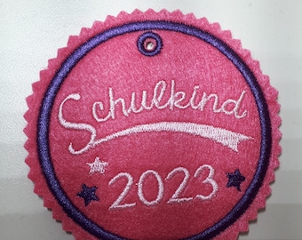 Op de afbeelding: Roze viltcirkel met een paarse rand en witte borduurwerk. De borduurwerk zegt "Schulkind 2023" met twee sterren.
