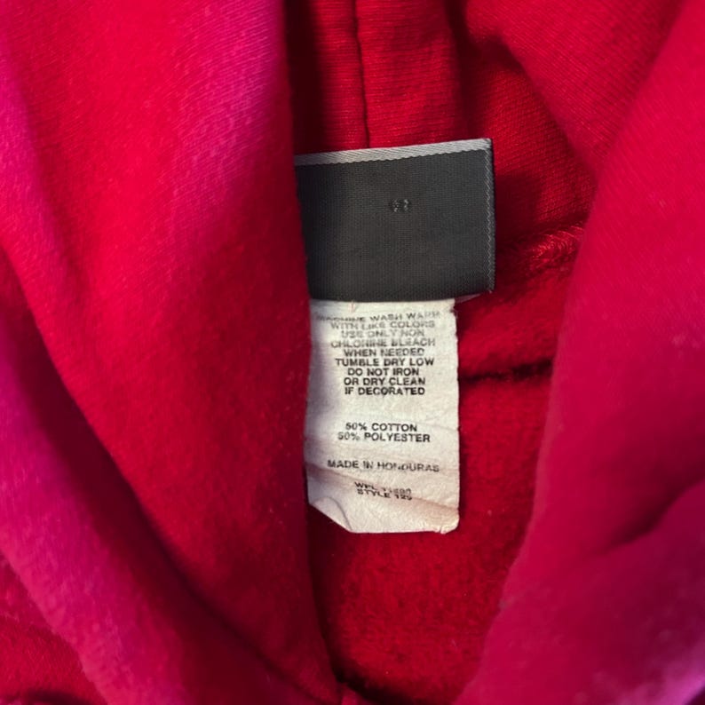 Puede incluir: Una sudadera con capucha de color rojo brillante que presenta una etiqueta de tela rectangular cosida en el interior del cuello. La etiqueta blanca muestra texto negro con instrucciones de cuidado, composici&oacute;n 50% algod&oacute;n y 50% poli&eacute;ster.
