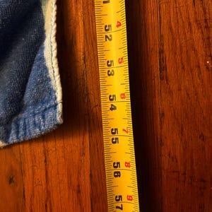 May include: A yellow tape measure with black markings, showing measurements from 132 to 145 cm. A blue and white fabric edge is visible on the left side. The tape measure is on a wooden surface.