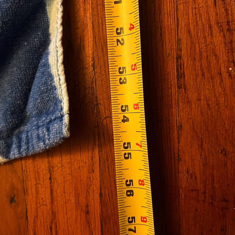 May include: A yellow tape measure with black markings, showing measurements from 132 to 145 cm. A blue and white fabric edge is visible on the left side. The tape measure is on a wooden surface.