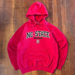 Puede incluir: Una sudadera con capucha de color rojo brillante que presenta el texto NC STATE en letras negras con bordes blancos, la palabra WOLFPACK en letras negras m&aacute;s peque&ntilde;as y un peque&ntilde;o logotipo NC debajo. La prenda incluye un bolsillo frontal y cordones.