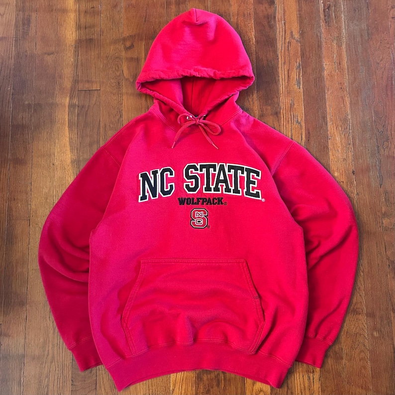 Puede incluir: Una sudadera con capucha de color rojo brillante que presenta el texto NC STATE en letras negras con bordes blancos, la palabra WOLFPACK en letras negras m&aacute;s peque&ntilde;as y un peque&ntilde;o logotipo NC debajo. La prenda incluye un bolsillo frontal y cordones.