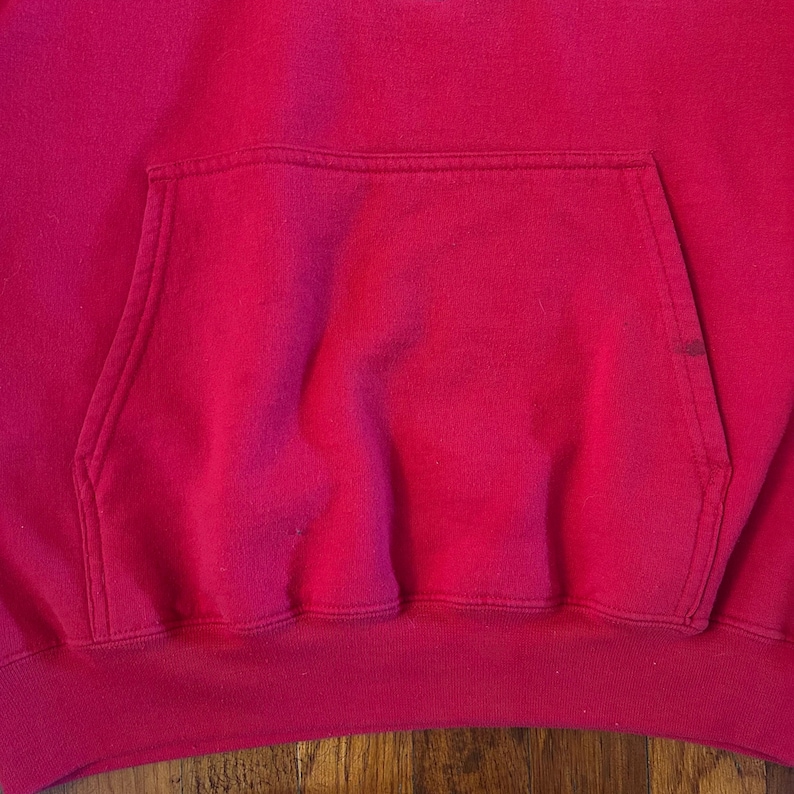 Puede incluir: Una sudadera con capucha de color rosa fucsia brillante que presenta un gran bolsillo canguro horizontal centrado en la parte delantera. La prenda est&aacute; confeccionada con un tejido suave y texturizado con costuras visibles en los bordes.