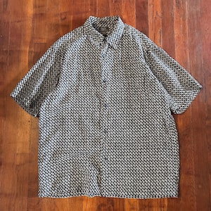 May include: A short-sleeved button-down shirt featuring a repeating black and white geometric triangle pattern. The shirt has a standard collar, a full-length front button placket, and a straight hemline, providing a consistent graphic design.