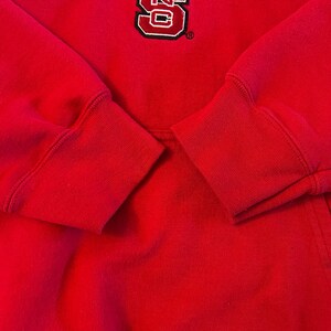 Puede incluir: Una sudadera roja de manga larga con un logotipo bordado prominente en el pecho. El logotipo consiste en una letra S roja grande y estilizada con un contorno blanco y borde negro, acompa&ntilde;ada por el texto NC State en letras blancas.