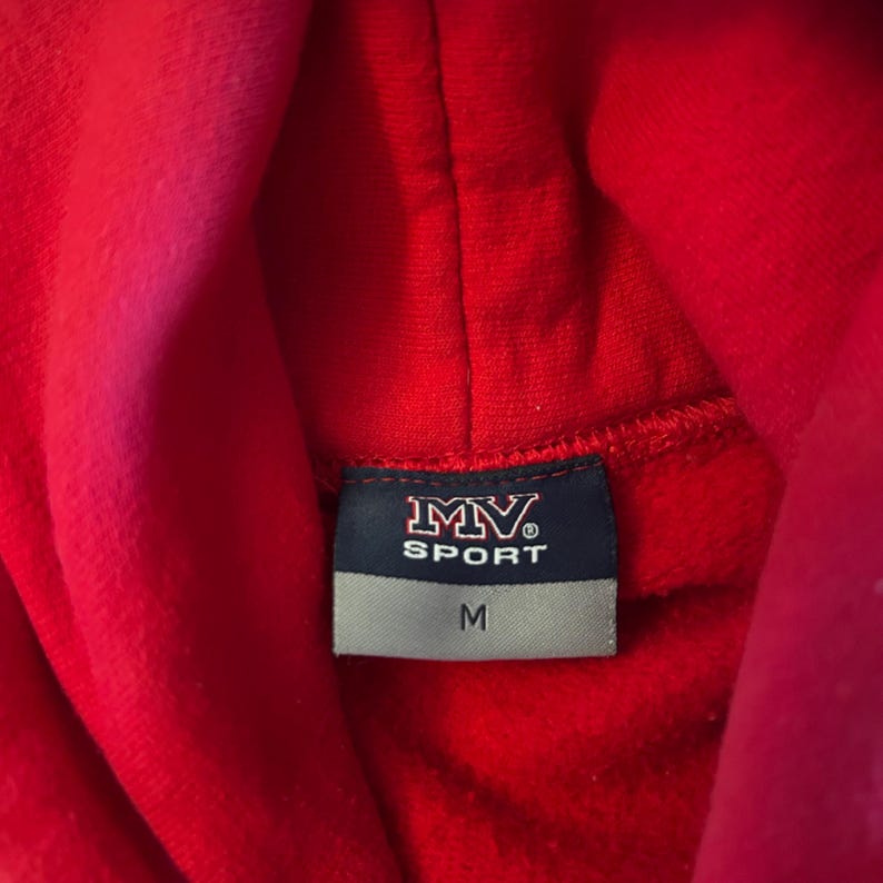 Puede incluir: Una sudadera con capucha de color rojo brillante que presenta una etiqueta rectangular en el cuello interior. La etiqueta tiene un fondo azul oscuro con el texto blanco MV SPORT y una secci&oacute;n inferior gris con la talla M en letras negras.